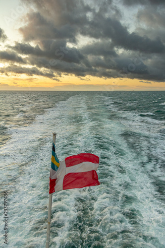 View from the stern of a ship at sunset with a flag and wake under a dramatic sky