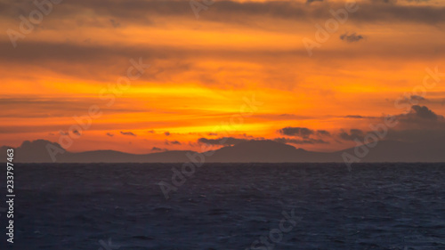 Brilliant orange and yellow sunset at sea with mountains and clouds in silhouette