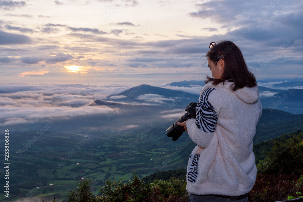 Fototapeta premium Woman tourist are looking at the camera after take a photo on nature landscape of the sun fog and mountains during the sunrise background at Phu Ruea National Park, Loei province, Thailand