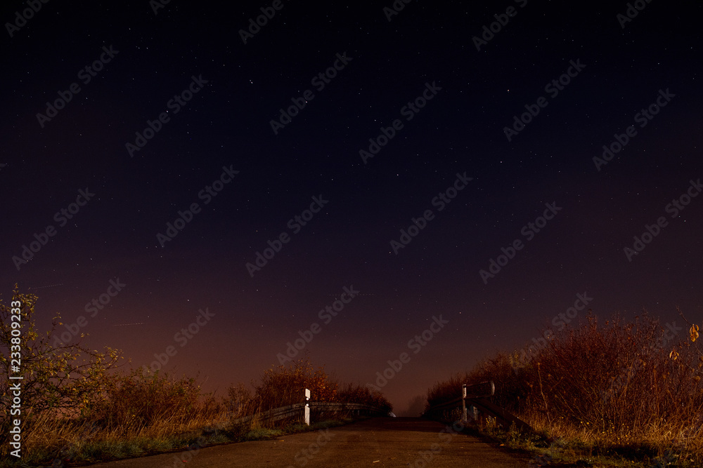 Countryside landscape view with a road leading to the nature at moody night with stars and blue night sky and mist glow. Braunschweig, Germany