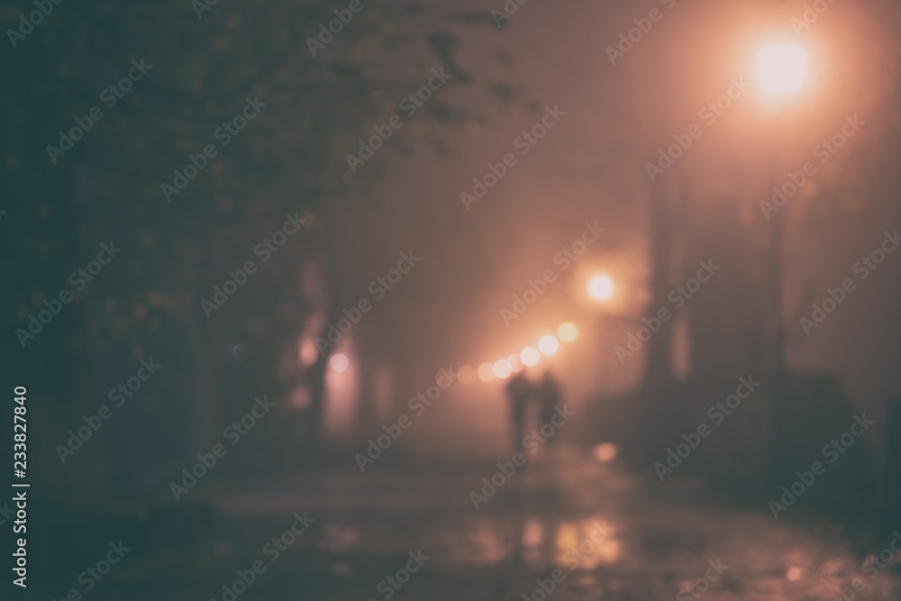 Foggy alley in night city park, blurred defocused background with silhouettes, burning lanterns, trees and benches