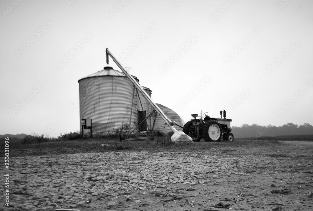 A black and white shot of a tractor, silo and grain auger in a field.