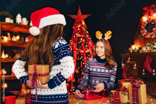 Cheerful mom wearing Christmas deer antlers and Rear view from the back of her daughter in santa hat holding in hands and exchanging gifts.