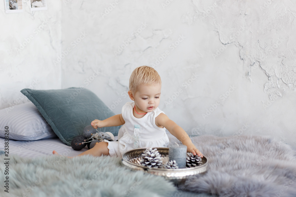 Fototapeta premium little baby girl in studio on bed. Beautiful little baby girl celebrates Christmas. New Year's holidays. Adorable baby girl playing with the garland on a gray background.