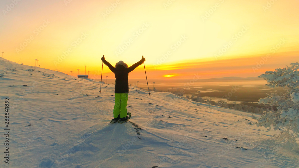 AERIAL Active woman snowshoeing snowy mountain slope and raising arms at the top