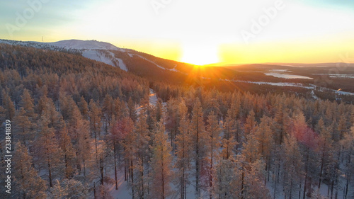 AERIAL: Gold winter sun setting behind frozen spruce forest and small ski resort