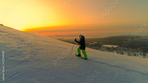 AERIAL: Active woman snowshoe trekking on snowy mountain slope at winter sunrise