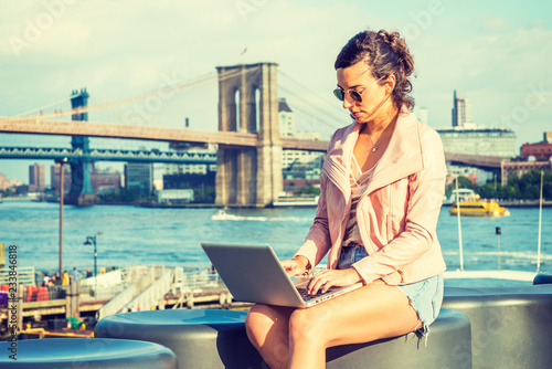 Young American Woman traveling, working in New York, wearing pink leather jacket, blue Denim shorts, sunglasses, sitting by river, working on laptop computer. Brooklyn, Manhattan bridges on background