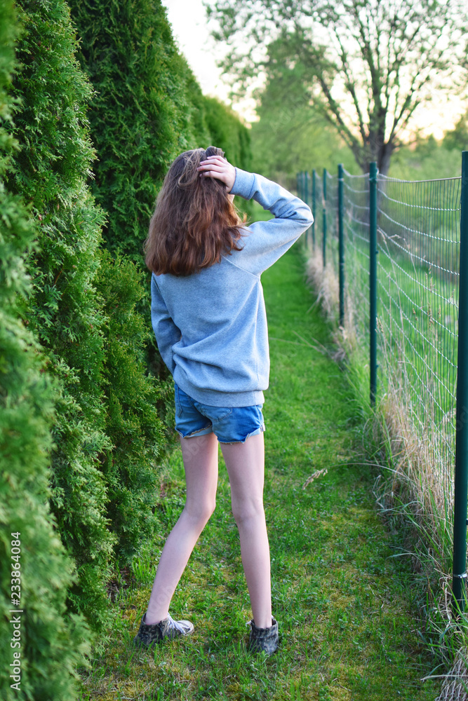 Teen girl wearing jeans shorts standing between green thuja hedge and fence  looking away in summer day back view. Stock Photo | Adobe Stock