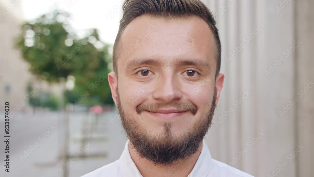 An attractive young man with a beard and wearing a white shirt smiling in town. Close-up shot. Soft focus An attractive young man with a beard and wearing a white shirt smiling in town. Close-up shot. Soft focus