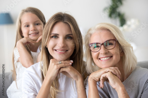 Portrait of happy three women generation, grandma mom and child granddaughter holding hands under chin looking at camera, young smiling adult daughter, older grandmother and kid girl family headshot photo