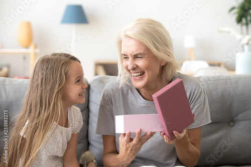 Happy mature grandmother opening gift box with present from grandchild girl laughing together, smiling granddaughter congratulating cheerful older grandma with birthday making pleasant fun surprise