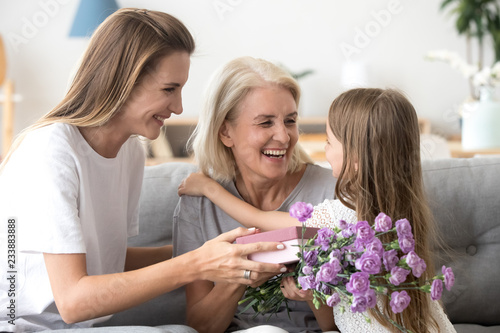 Happy grandma embracing little granddaughter thanking grandchild and grown daughter for gift box holding flower bouquet, smiling child girl and mother congratulate granny with birthday giving present