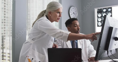 Portrait of millennial black doctor using computer while senior colleague directs him. Two doctors working inside medical office looking at computer monitor and typing