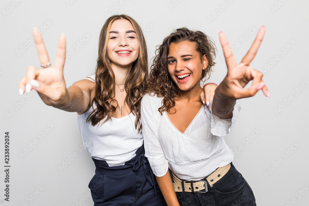 Two happy pretty women posing together and showing peace gestures while looking at the camera over gray background Two happy pretty women posing together and showing peace gestures while looking at the camera over gray background