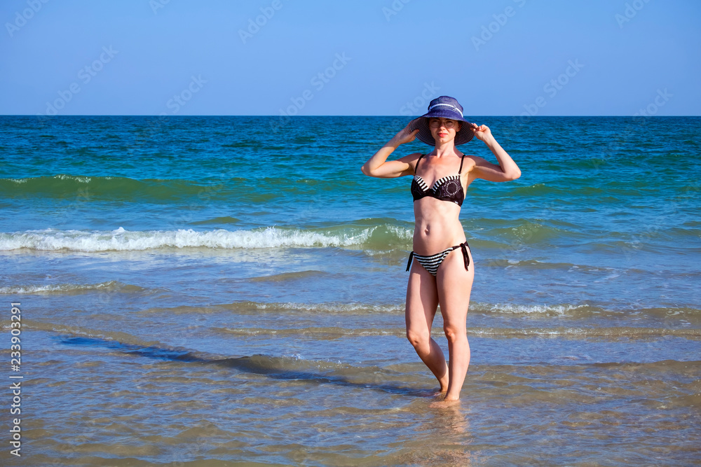 Young beautiful girl in a bathing suit and a striped hat is standing in the sea with her hands on the brim hat Young beautiful girl in a bathing suit and a striped hat is standing in the sea with her hands on the brim hat