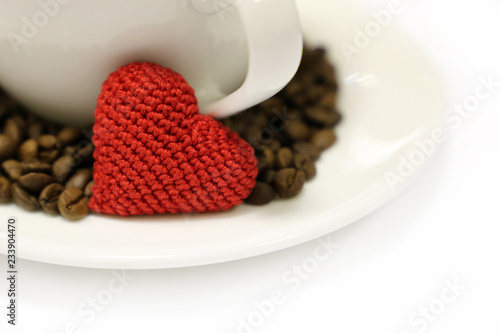 Coffee cup, red knitted heart and coffee beans on white saucer. Concept of romantic breakfast, love to coffee, Valentine's day card