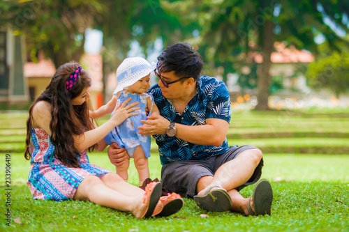 young happy loving Asian Korean parents couple enjoying together sweet daughter baby girl sitting on grass at green city park in love and parenthood concept