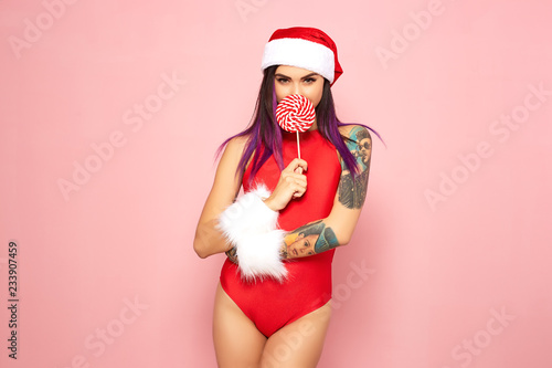 Girl with purple hair tips and tattoo on her arm dressed in red swimsuit, Santa's hat and white fur bracelets holding a lollipop in front of her face on the background of pink wall