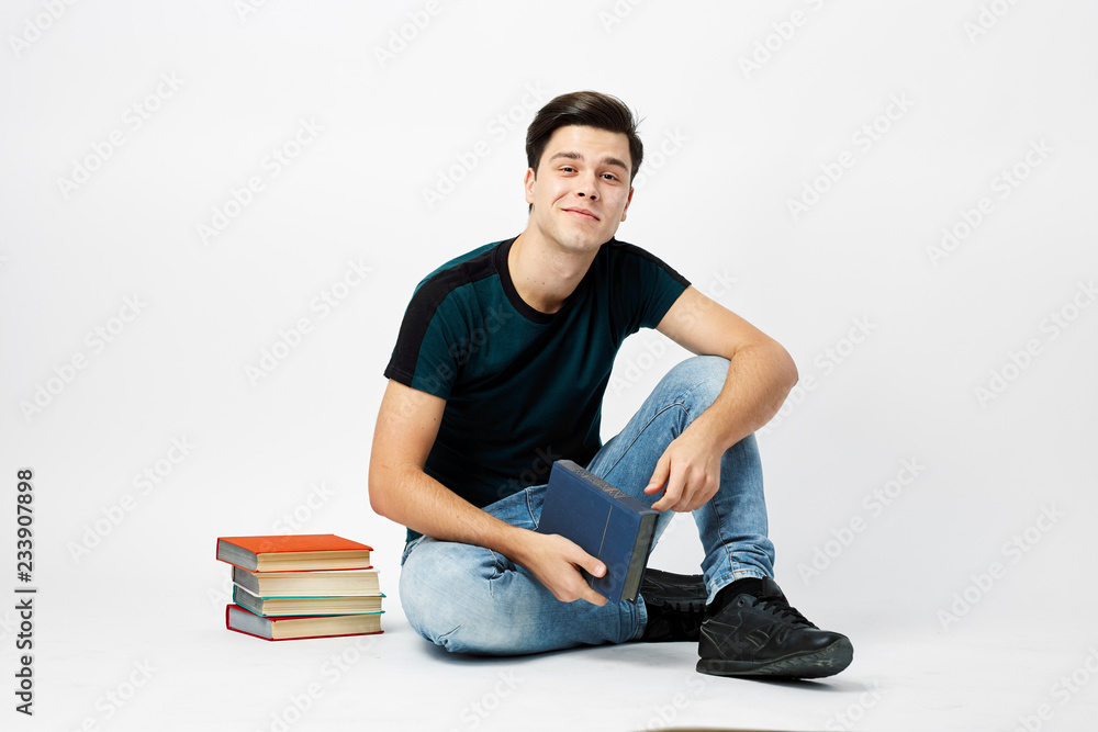 Nice dark-haired guy dressed in a dark t-shirt and jeans sits on the floor and reads a book on a white background in the studio