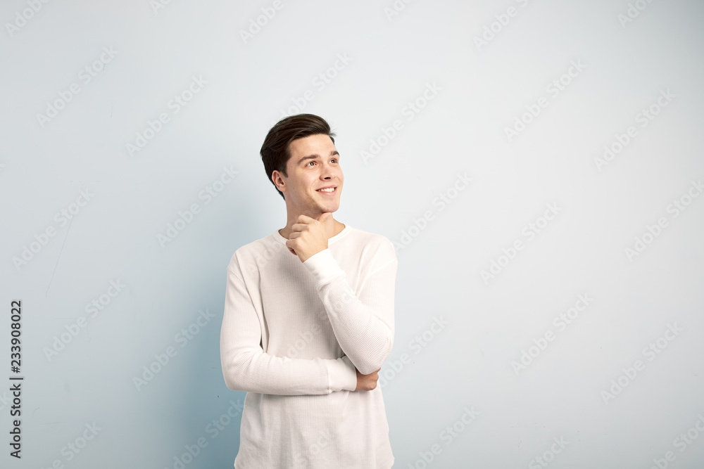 Smiling dark-haired guy dressed in a white long sleeve t-shirt and jeans stands and keeps his hand on his chin on a white background Smiling dark-haired guy dressed in a white long sleeve t-shirt and jeans stands and keeps his hand on his chin on a white background