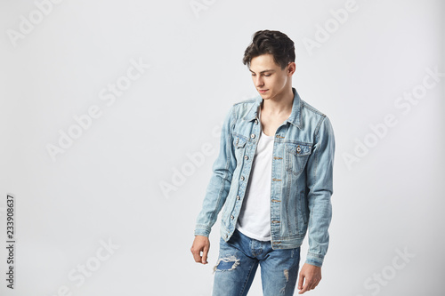 Young dark-haired guy in a white t-shirt, jeans and a denim jacket stands on the white background in the studio