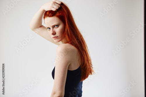 Charming redhead long-haired girl dressed in black top and jeans holds hand on her head on the white background in the studio