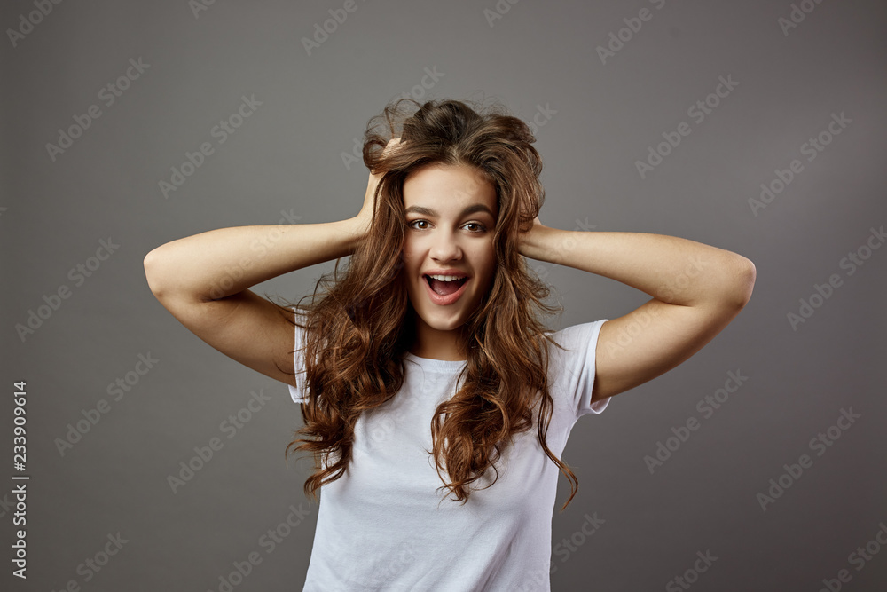 Fototapeta premium Funny girl with long brown hair dressed in a white t-shirt holds her hands on her head in the studio on the gray background