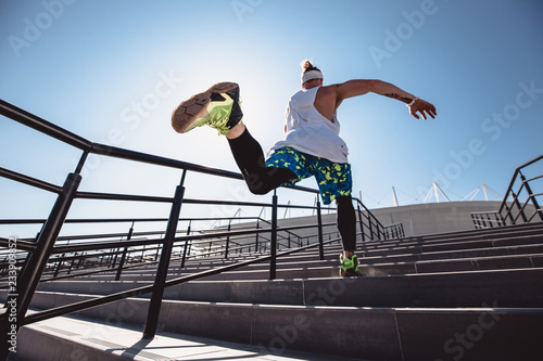 Athletic young man with headband on his head dressed in the white t-shirt, black leggings and blue shorts is jumping up the stairs outside on a sunny day