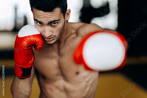 Sportsman with a naked torso and the red boxing gloves his hands stands hits with left hand in the boxing gym