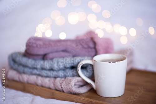 Cup of coffee with stack of knitted clothes over Christmas lights. Winter season.