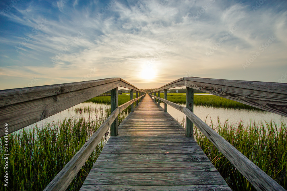 coastal waters with a very long wooden boardwalk pier in the center during a colorful summer sunset under an expressive sky with reflections in the water and marsh grass in the foreground