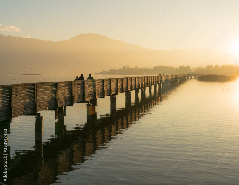 long wooden pier and boardwalk over Lake Zurich near rapperswill in golden evening light with silhouette of pedestrians and people walking and mountains in the background