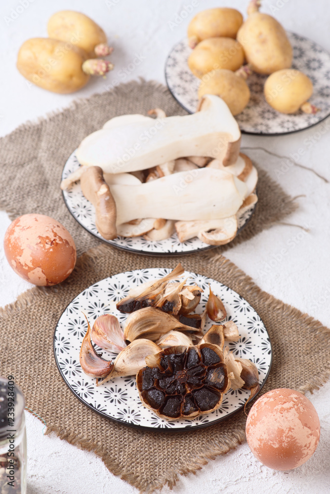 Fototapeta premium Black garlics eggs potatoes and other raw ingredients on textured white base