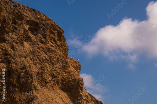 Rugged jagged brown cliff edge with a seagull on a ledge. Deep blue sky background with broken puffy clouds