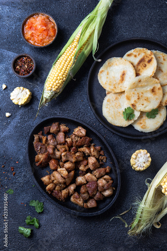 COLOMBIAN FOOD. Maize AREPAS and fried pork chicharron ans colombian tomato sauce. Top view. Black background