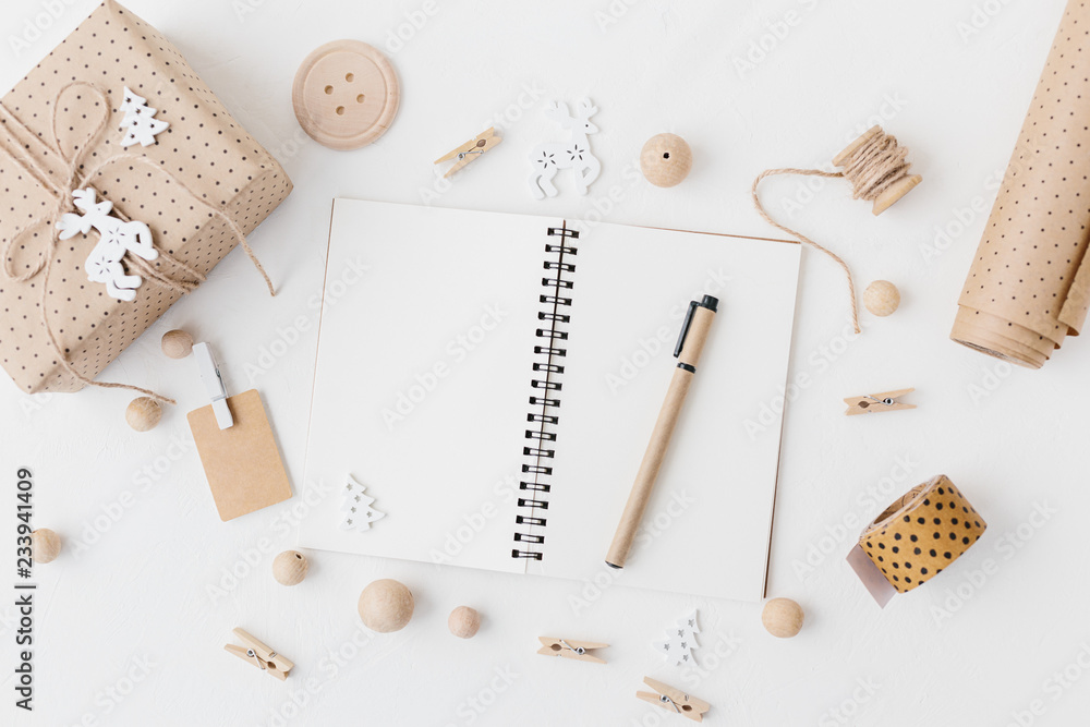Open notebook, pen, wrapped craft gift, wrapping paper, gift label, wooden beads on textured white background. Open notebook, pen, wrapped craft gift, wrapping paper, gift label, wooden beads on textured white background.