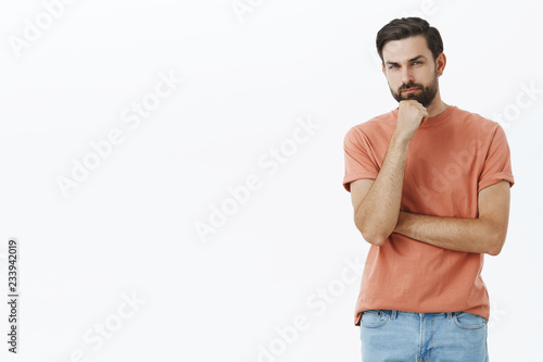 Guy having hesitations, thinking. Portrait of unsure and thoughtful attractive nice bearded guy with dark hair squinting doubtful and uncertain holding fist on chin as making choice over gray wall © Cookie Studio