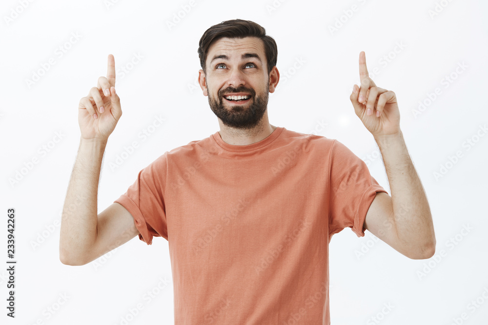Fototapeta premium Happy curious attractive european man with dark beard in casual t-shirt raising index fingers looking and pointing up wondered and delighted enjoying nice view on stars over gray background