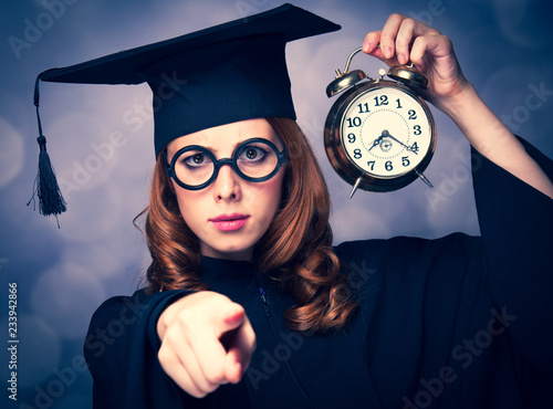 Redhead student girl in cap and gown holding a metal classic alarm clock on green background with bokeh