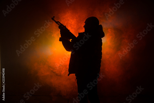 Fototapeta Naklejka Na Ścianę i Meble -  Silhouette of man with assault rifle ready to attack on dark toned foggy background or dangerous bandit in black wearing balaclava and holding gun in hand. Shooting terrorist with weapon theme decor