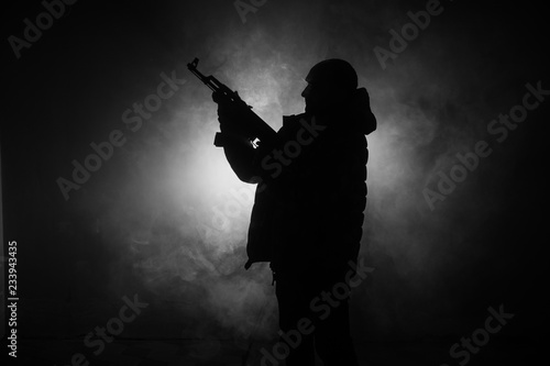 Silhouette of man with assault rifle ready to attack on dark toned foggy background or dangerous bandit in black wearing balaclava and holding gun in hand. Shooting terrorist with weapon theme decor