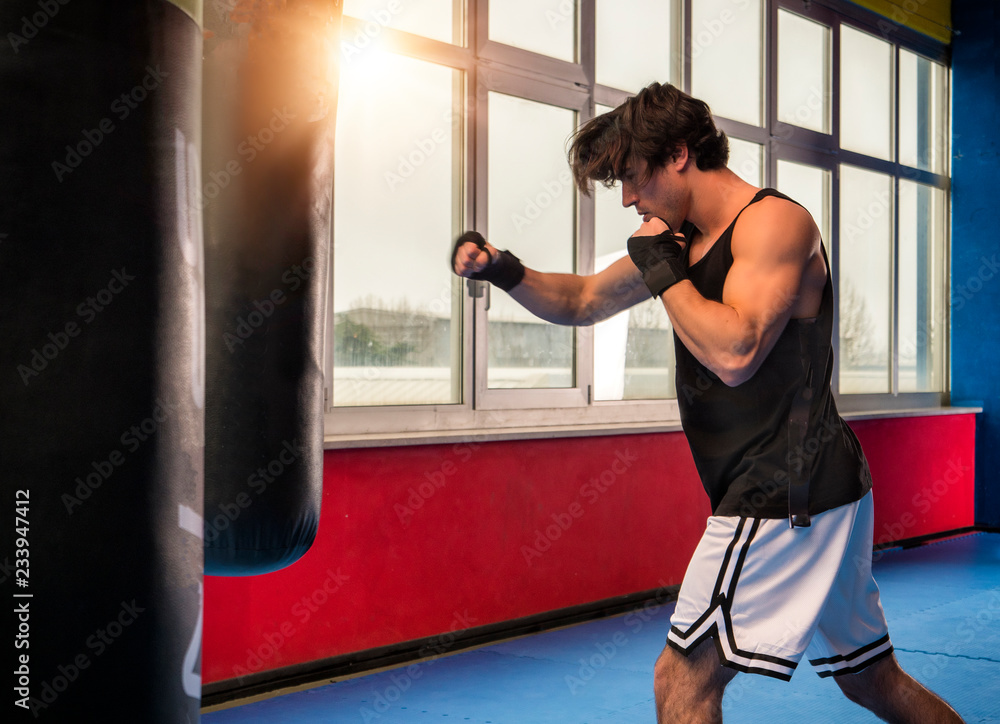 Side view of muscular sportive man in modern gym training with punching bag while boxing alone in back lit