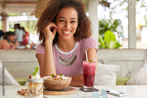 Photo of attractive black woman has Afro hairstyle  dressed in casual t shirt  smiles broadly at camera  rejoices meeting with colleague in cozy cafe  drinks fresh smoothie  likes exotic food