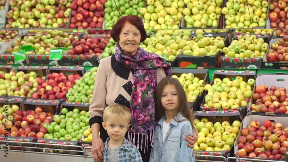 Happy cheerful elderly woman in her grandchildren to buy food and fruit at the supermarket. Portrait of grandmother and grandchildren on the background of boxes of apples in the grocery store. Happy cheerful elderly woman in her grandchildren to buy food and fruit at the supermarket. Portrait of grandmother and grandchildren on the background of boxes of apples in the grocery store.