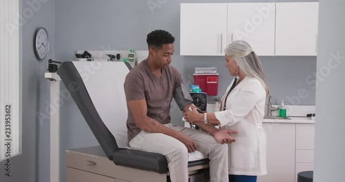 Mature female medical practitioner measuring blood pressure with monitor and stethoscope of a young male patient. Attractive black man having his blood pressure measured by senior white doctor