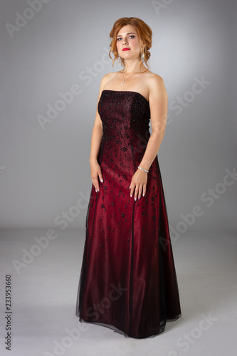 Luxurious young woman in a long red evening dress on a gray background in full height.