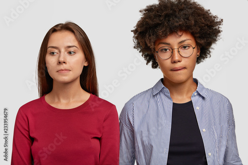 Indoor shot of contemplative mixed race women purse lips, being deep in thoughts, have sad expressions, dressed in casual clothes, isolated over white background. Facial expressions concept.