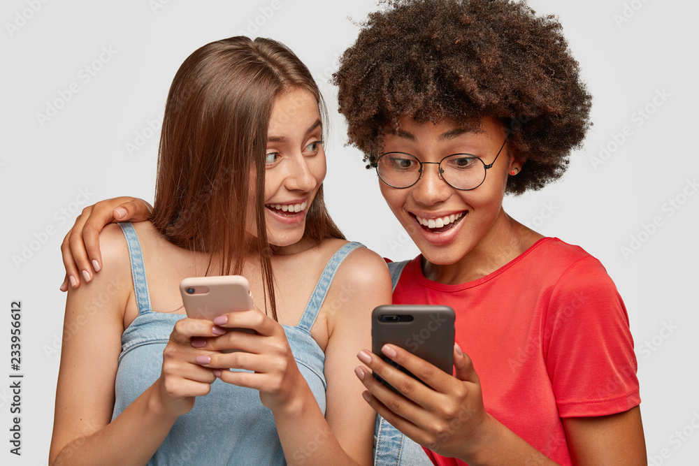 Multiracial friends embrace and share multimedia files via cell phone bluetooth, enjoy wireless connection together, look gladfully at cellulars, isolated over white background. Friendship concept Multiracial friends embrace and share multimedia files via cell phone bluetooth, enjoy wireless connection together, look gladfully at cellulars, isolated over white background. Friendship concept