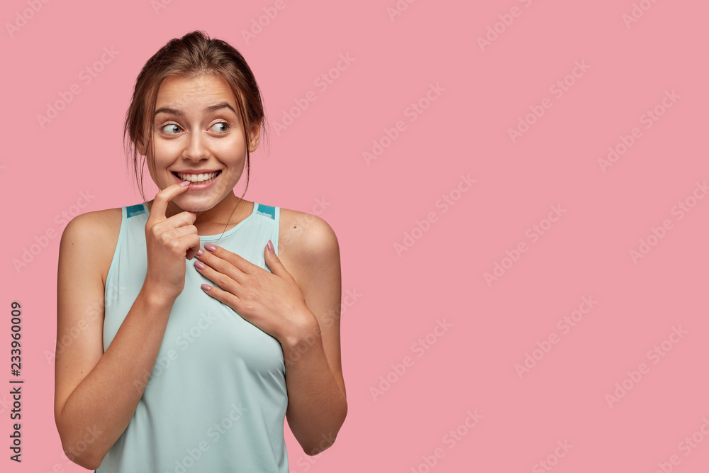 Isolated shot of positive European woman bites finger, keeps hand on chest, smiles positively, looks aside, wears casual clothes, hears intriguing information, isolated over pink wall with free space Isolated shot of positive European woman bites finger, keeps hand on chest, smiles positively, looks aside, wears casual clothes, hears intriguing information, isolated over pink wall with free space